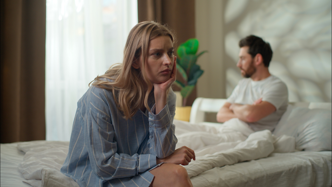 Frau wirkt nachdenklich und belastet, während Partner im Hintergrund distanziert sitzt